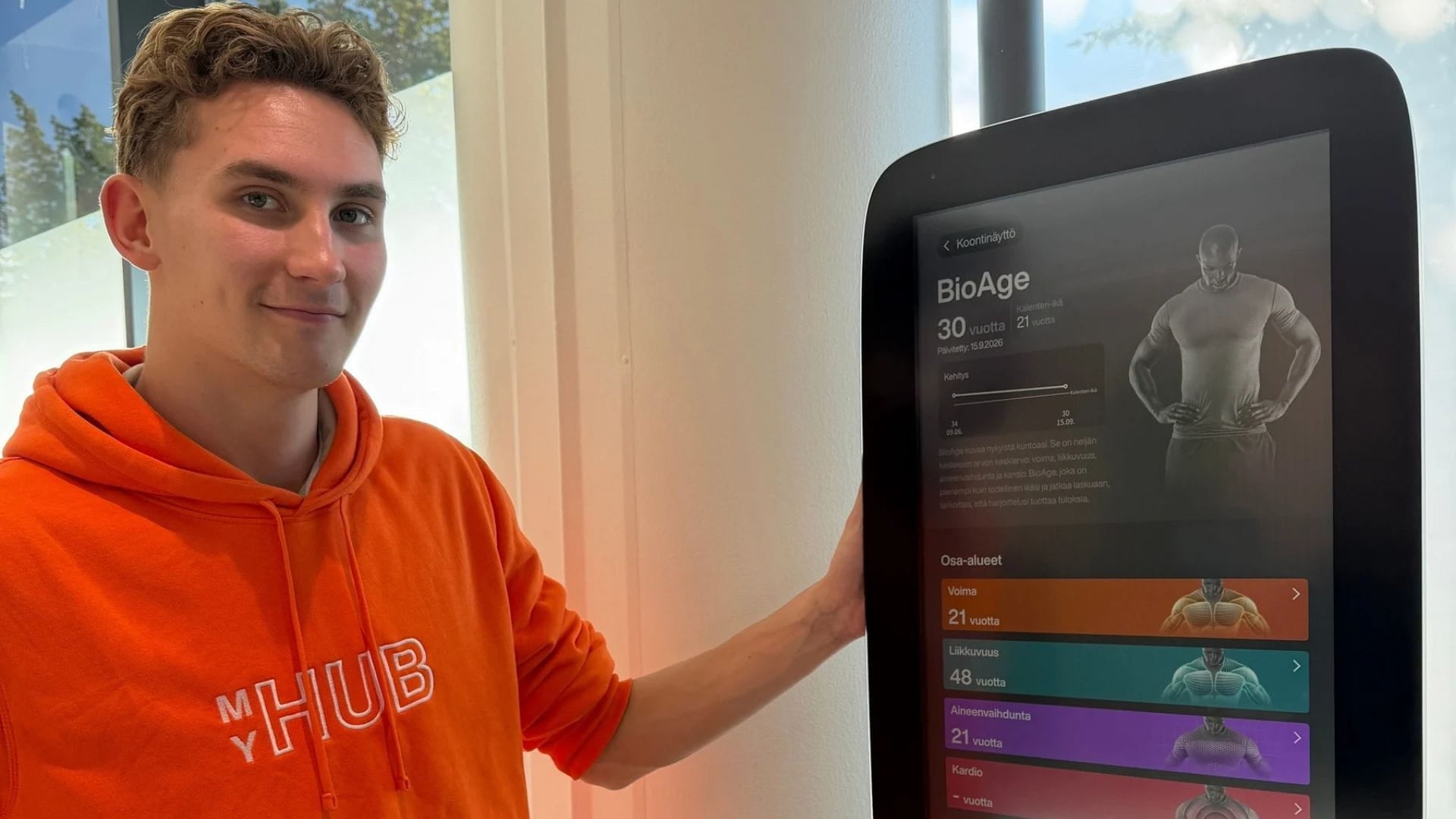 Kuvassa mies oranssissa MyHub-hupparissa poseeraa oman bio-ikä tuloksensa kanssa. 