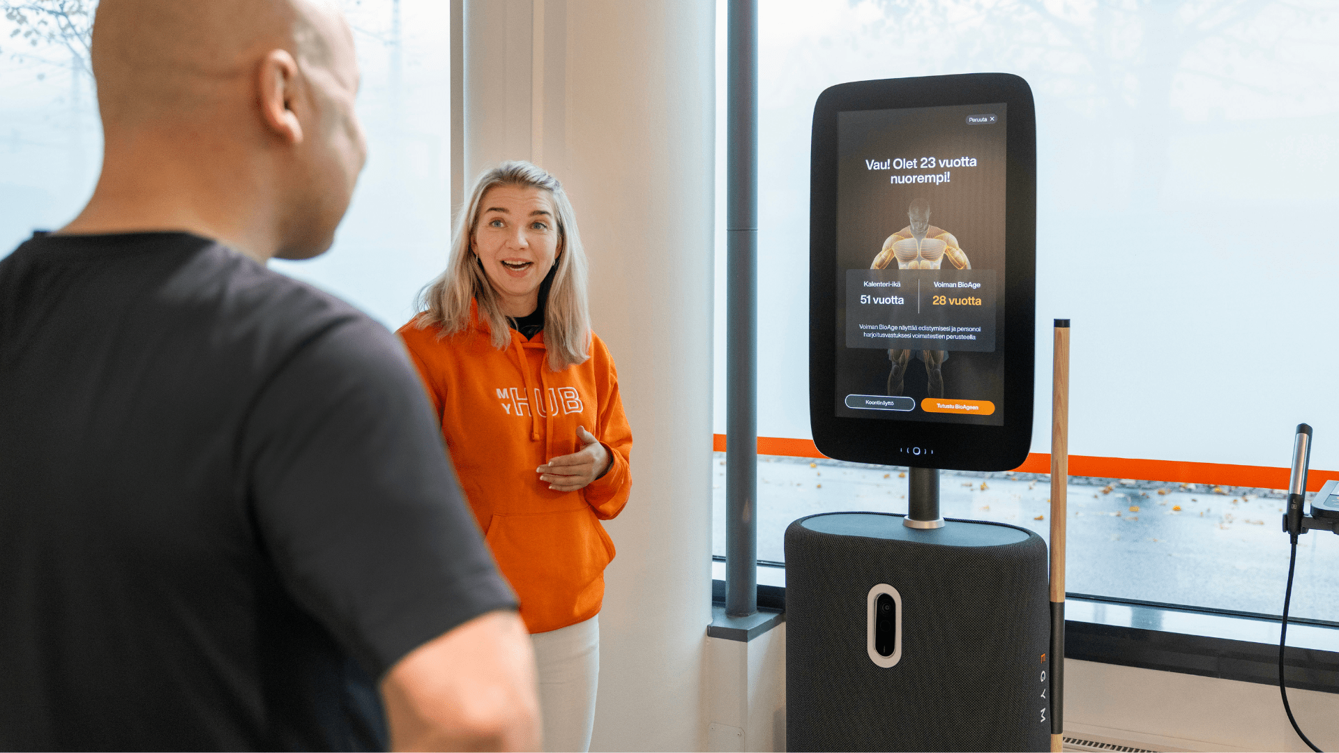 MyHub Fitness Hub kertoo bio-iän miehelle. Näytöllä lukee: Vau! Olet 23 vuotta nuorempi! MyHub host seisoo näytön vieressä iloisena.